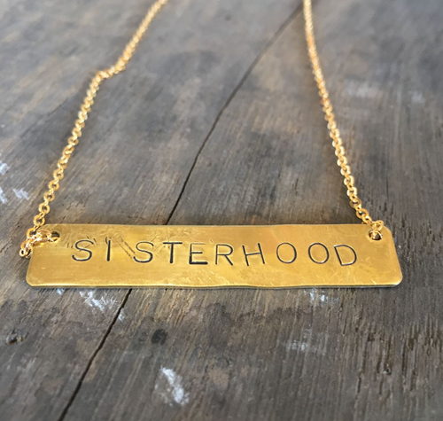 A bar necklace with the word SISTERHOOD  stamped into the brass. It hangs on a gold plated chain 16 inches long.