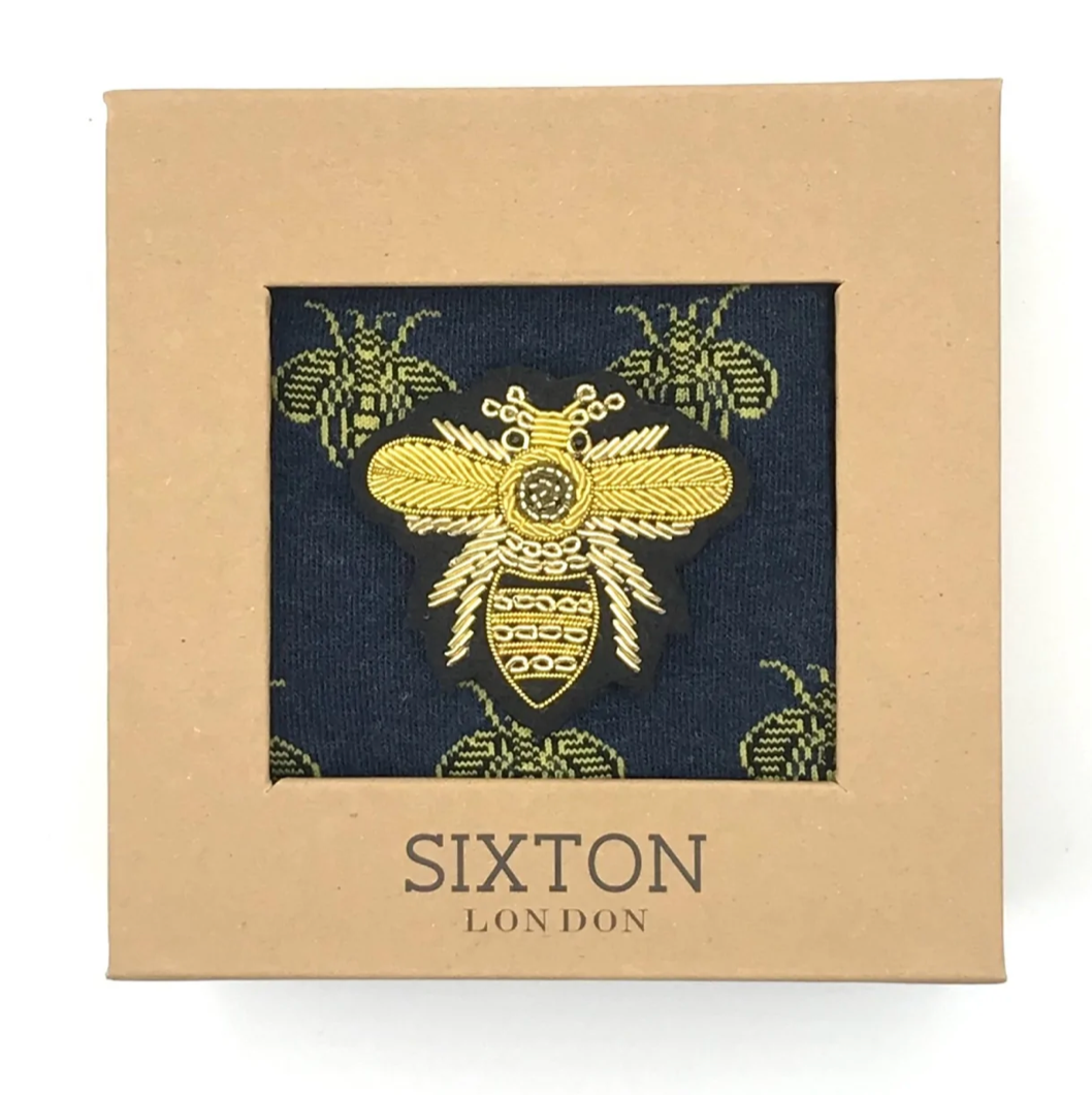 A pair of navy blue socks with yellow bee designs on them and a large bee pin. All presented in a box.