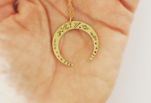 A brass crescent moon pendant necklace with hammered mystic designs hammered into it.