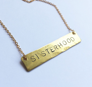 A bar necklace with the word SISTERHOOD  stamped into the brass. It hangs on a gold plated chain 16 inches long.