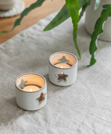 A pair of small white ceramic tea light holders with stars cut out of them allowing the light to shine through