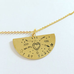 A semi circle brass pendant hand stamped with a heart and various symbols with the words I AM MAGIC on a gold plated chain