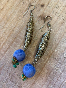 Handmade and fairtrade earrings using gold metal intricately wound round to create a drop pendant. To the base is a blue fabric covered bead, covered using material from recycled sari's. To the very base are tiny green beads which dangle and move as you do. 